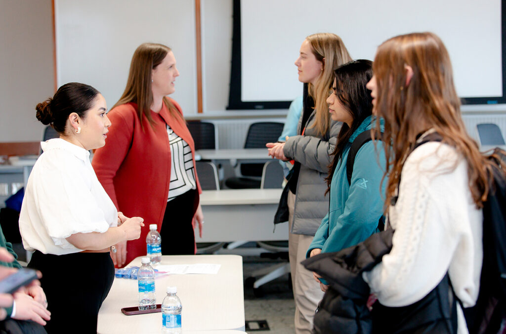 Students speak with two women presenters during a networking session in a university classroom. The students are standing across from a table with water bottles and informational materials, while additional conversations take place in the background.