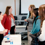 Students speak with two women presenters during a networking session in a university classroom. The students are standing across from a table with water bottles and informational materials, while additional conversations take place in the background.