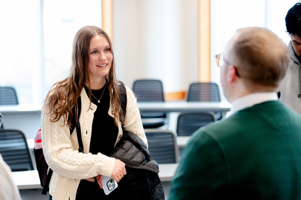 A student wearing a cream cable-knit cardigan, black shirt, and backpack smiles while speaking with two people in a bright university classroom. She holds a phone and a black jacket as she engages in conversation, with rows of desks and chairs softly blurred in the background.