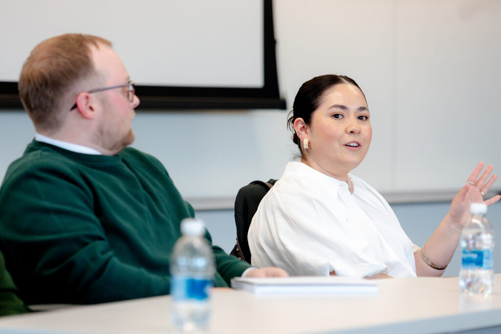 A man in a great sweater and a woman in a white blouse sit at a table during a networking session in a university classroom.