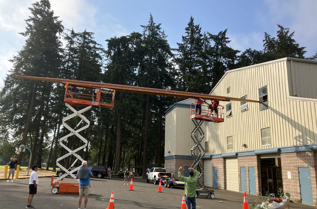 Workers use two scissor lifts to guide a long cedar racing shell out of a boathouse.
