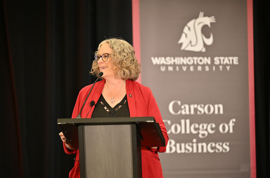 A person wearing a red jacket over a black top speaks at a podium while standing in front of a Washington State University Carson College of Business banner at a formal event.
