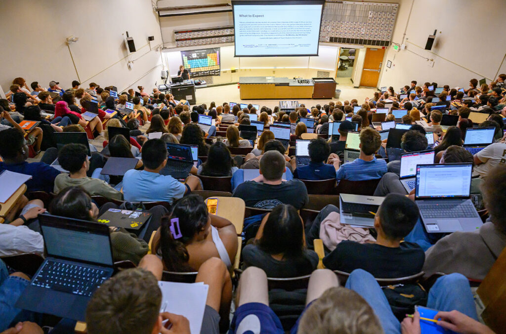 A room full of students in a lecture hall watch as a professor lectures in front of a screen.