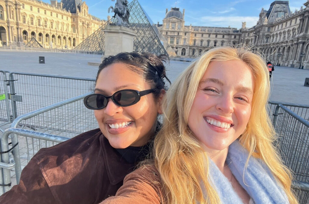 Two people (Scoggins left) take a selfie in the courtyard of the Louvre. The Louvre Pyramid and ornate historic museum buildings are visible behind them, along with metal crowd-control barriers under a clear blue sky.