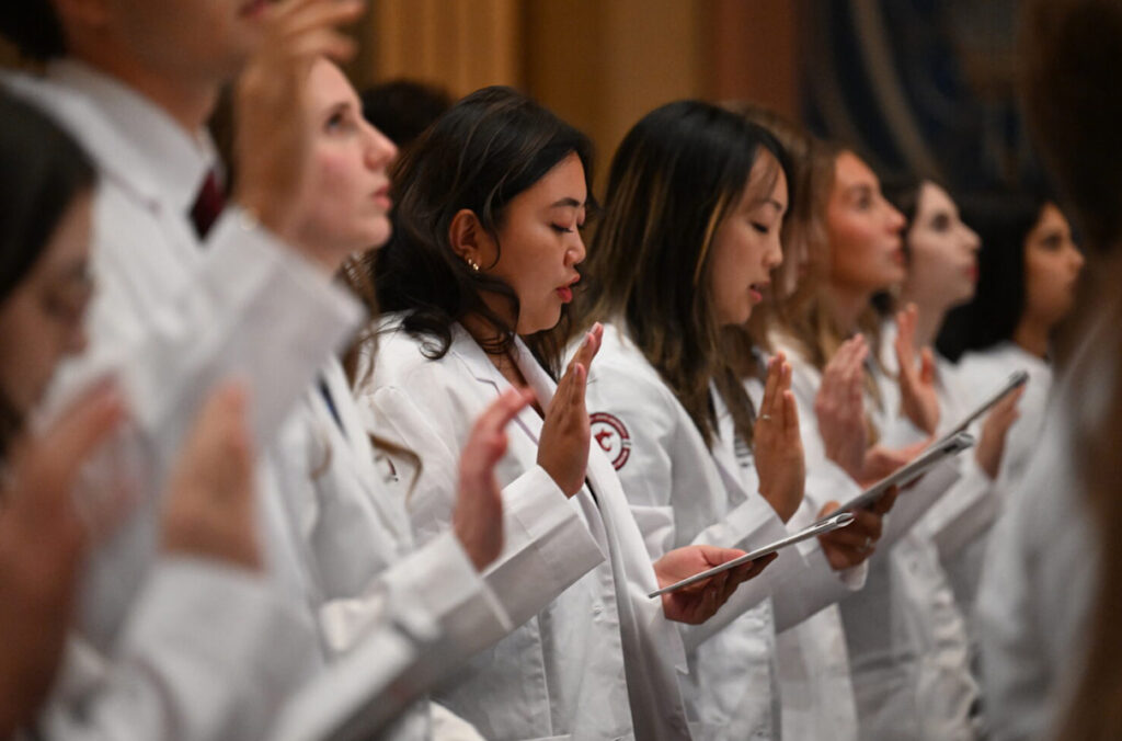 Medical students in white coats stand in a row with raised hands and printed programs, reciting a pledge during a White Coat Ceremony.