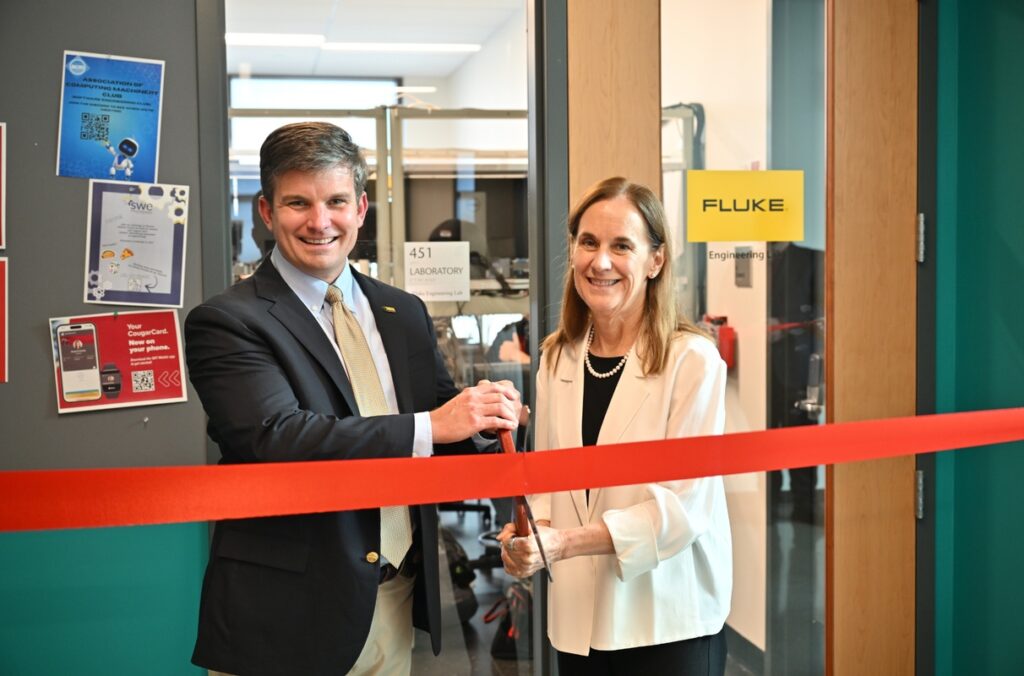 Two people stand behind a red ribbon during a ribbon-cutting ceremony inside an engineering laboratory. One person holds large ceremonial scissors while both stand in front of a glass-walled lab entrance. A yellow "FLUKE" sign and laboratory equipment are visible in the background.