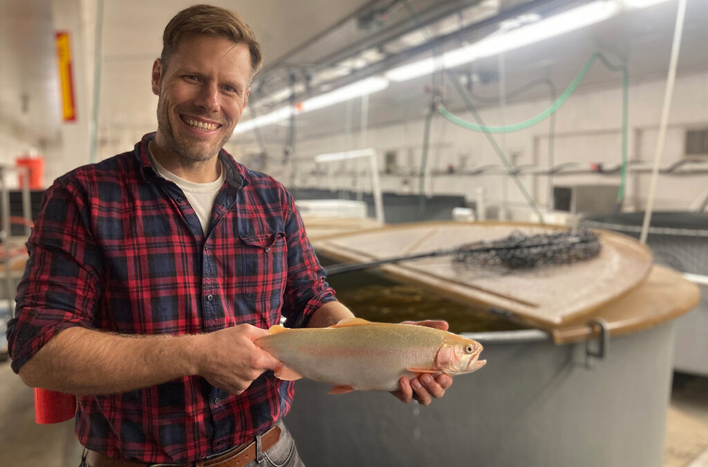 Phelps, holding a large pink-colored trout in a long room lined with pond-like tanks, conduits, and lights. A fish net rests on a nearby tank.