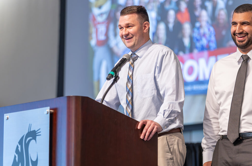 Family medicine residency graduate Dr. Jeff Ward speaks at a podium bearing the Washington State University logo while Dr. Mohammed Younes stands nearby during a graduation ceremony.
