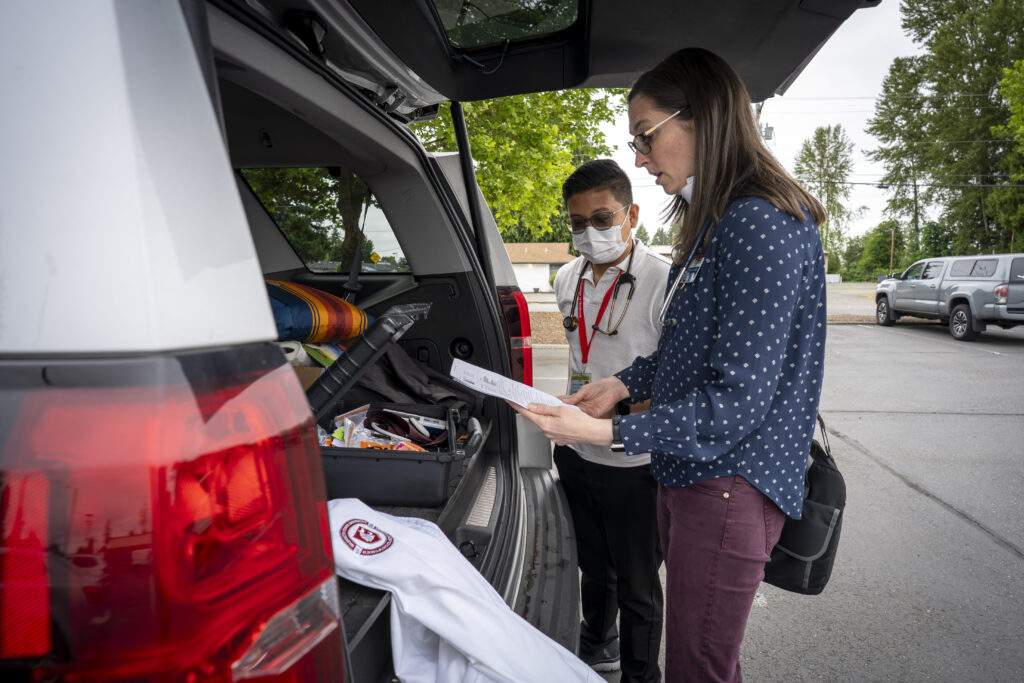 Healthcare team members review a checklist and medical supplies in the open rear cargo area of a mobile health vehicle parked outdoors.
