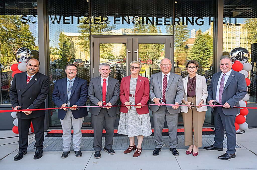 Ribbon-cutting ceremony outside the Schweitzer Engineering Hall entrance, with seven individuals standing in front of the building holding a red ribbon and ceremonial scissors. Red, white, and gray balloon columns flank the entrance, and the building’s glass façade reflects surrounding campus scenery.