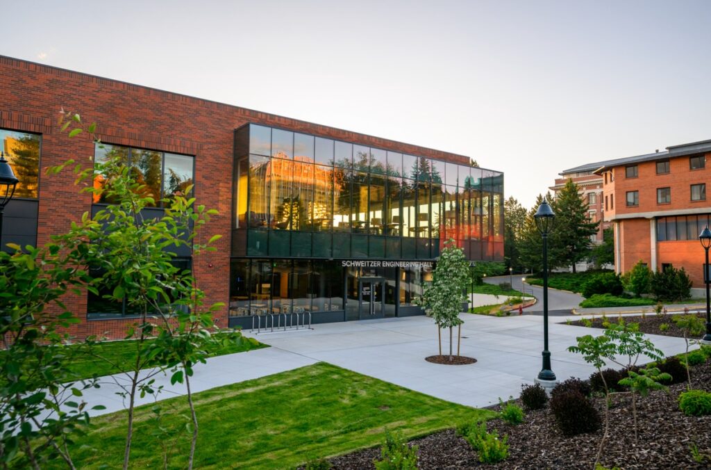 Exterior view of Schweitzer Engineering Hall, a modern red-brick academic building with a prominent glass facade reflecting the surrounding campus. The foreground features landscaped lawns, trees, and paved walkways, while additional campus buildings and lamp posts are visible in the background under soft evening light.
