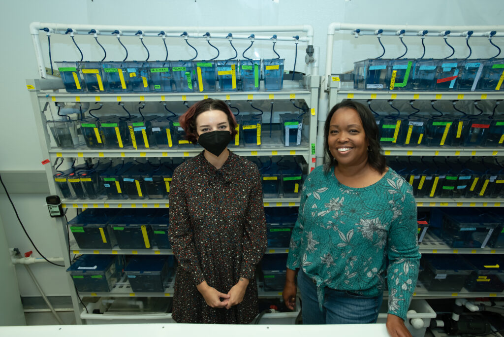 Two women pose for a picture in front of tanks of zebrafish.