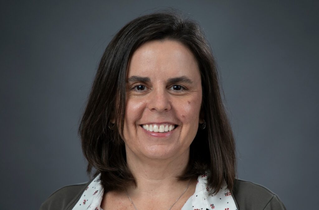 Professional studio portrait of a person with shoulder-length dark hair, wearing a gray jacket over a white patterned blouse and a necklace, centered against a gray gradient background.