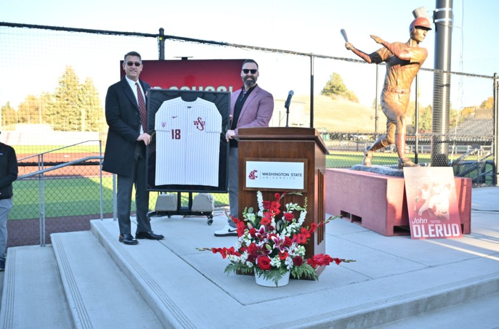 Two individuals stand on an outdoor stage displaying a framed Washington State University baseball jersey with the number 18. A podium bearing the Washington State University logo and a red-and-white floral arrangement are in the foreground. A large bronze statue of a baseball player stands to the right beside a sign reading ‘John Olerud,’ with the baseball field and stadium seating visible in the background.”