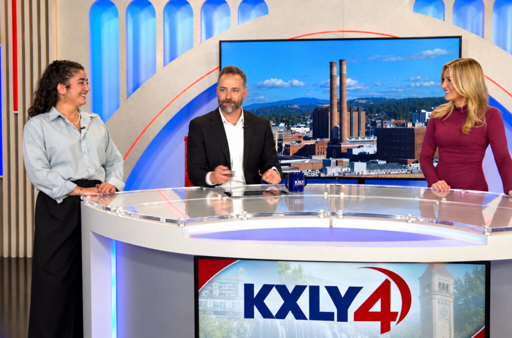 Armenian journalism student Lori Ishilian stands next to two newscasters at a desk who are giving a live broadcast.