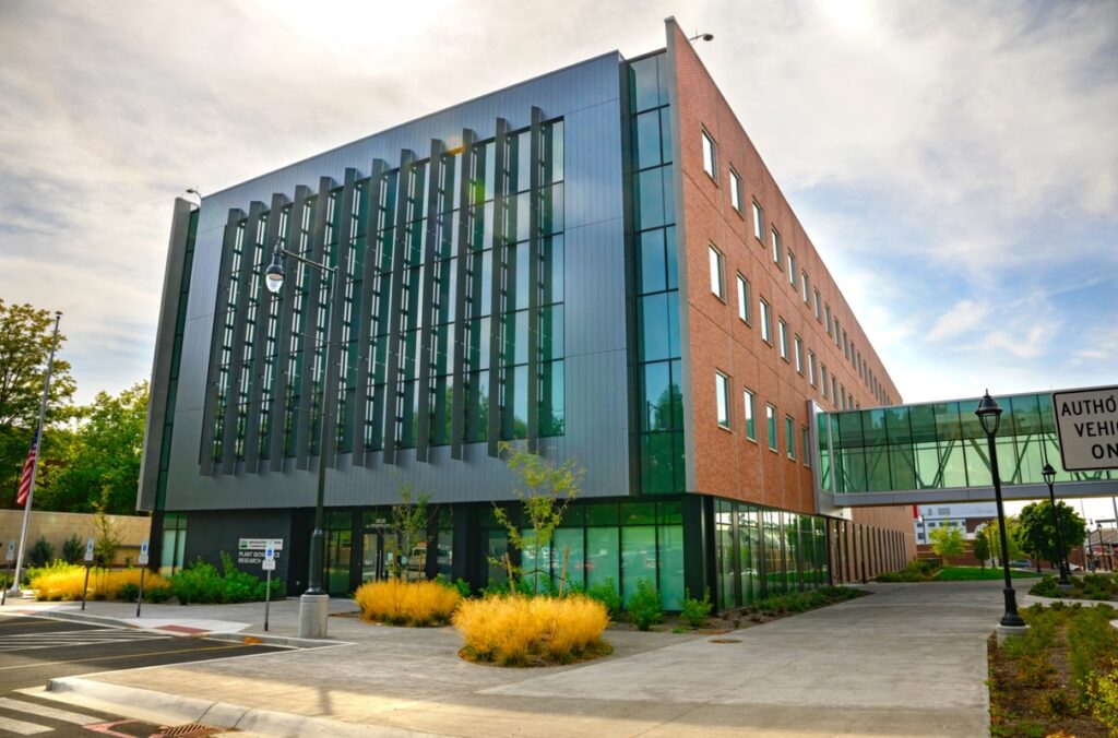 Modern research facility with a dark metal-and-glass façade and adjoining red-brick wing, viewed from a street corner. Landscaped plantings and sidewalks surround the building, and an enclosed glass skybridge connects to another structure under a partly cloudy sky.