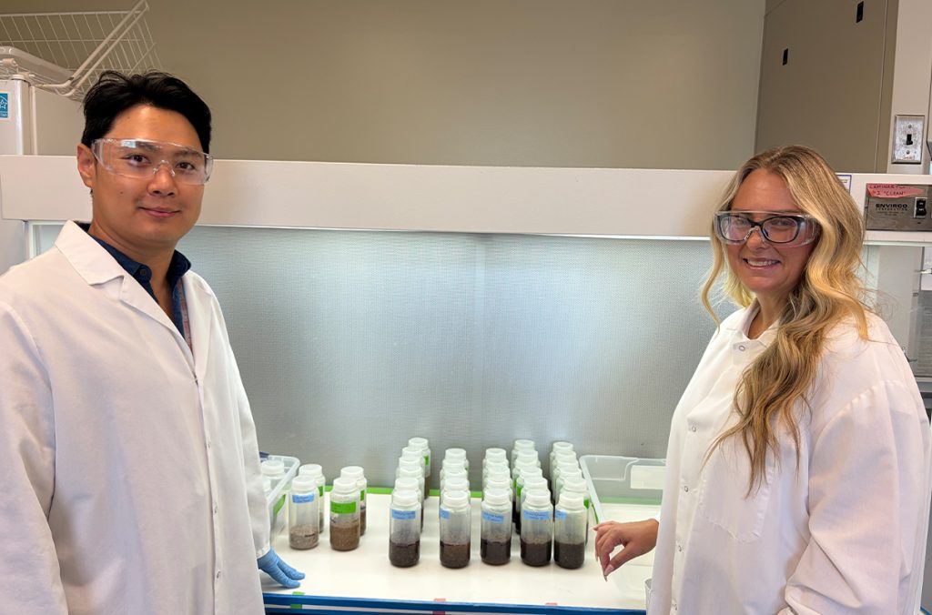 Two researchers in a laboratory stand next to a fume hood displaying rows of sample bottles containing agricultural or biomass materials being used in a research project.