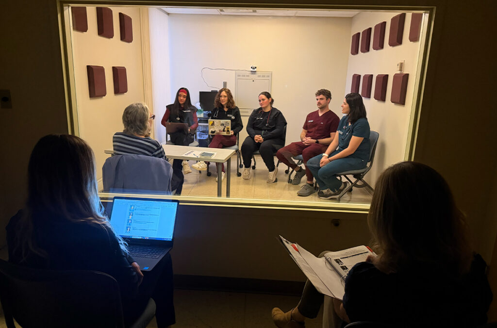 Two people with laptops watch a group of veterinary medicine students and actors perform a mock patient visit through a window.