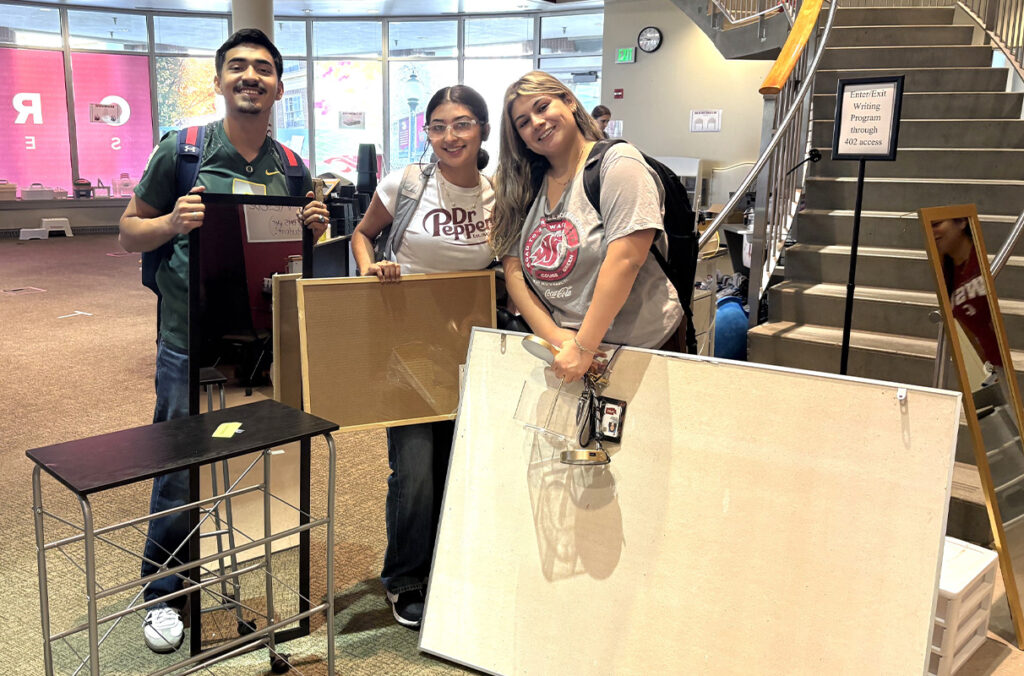 Three students pose for a picture with used items in the atrium of a residence hall.
