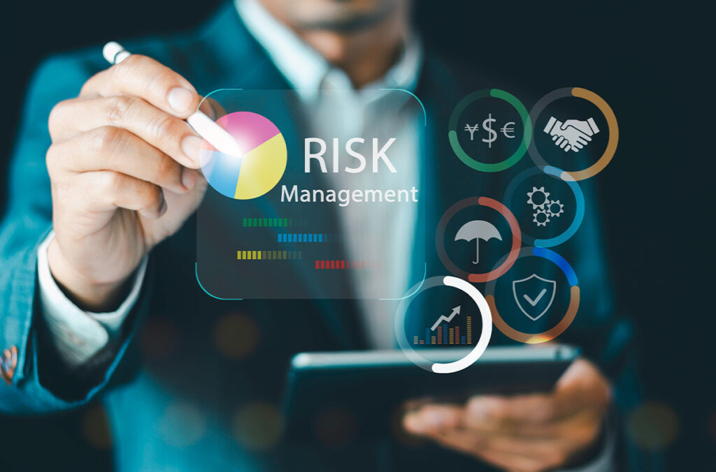 A person in a business suit uses a stylus on a tablet while a transparent digital interface displays “Risk Management,” a colorful pie chart, and icons such as currency symbols, a handshake, an umbrella, gears, a shield, and a bar chart.