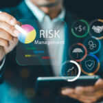 A person in a business suit uses a stylus on a tablet while a transparent digital interface displays “Risk Management,” a colorful pie chart, and icons such as currency symbols, a handshake, an umbrella, gears, a shield, and a bar chart.