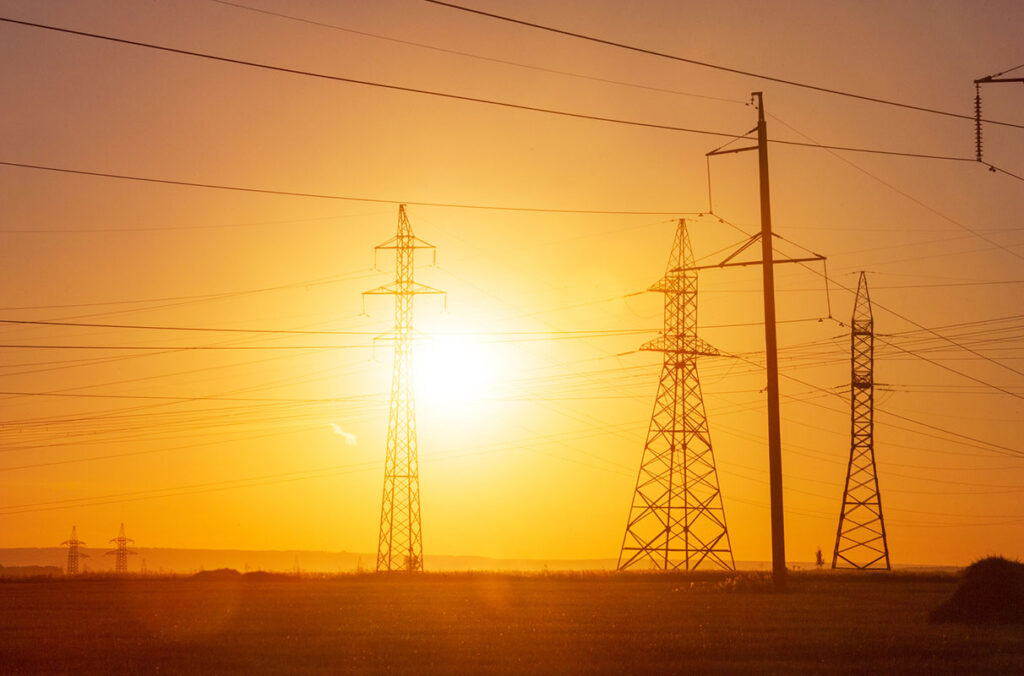 High-voltage power transmission lines and towers against an orange sky with a bright glowing un in the background.