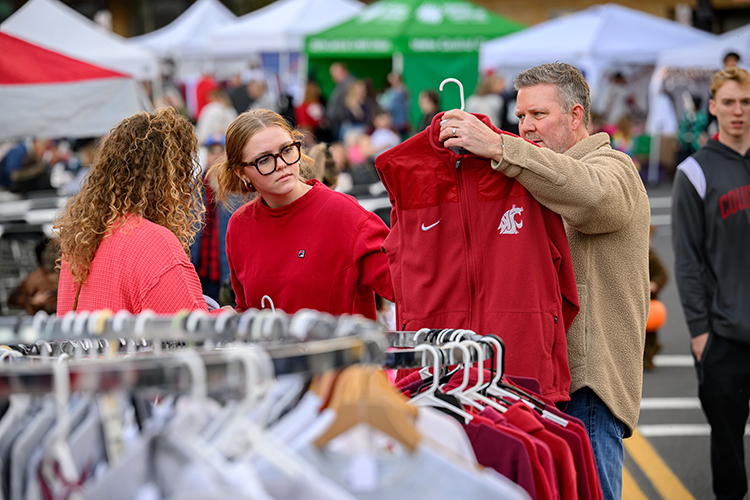 A father holds up a WSU jacket for sale to show his family. Other vendor booths are visible in the background.