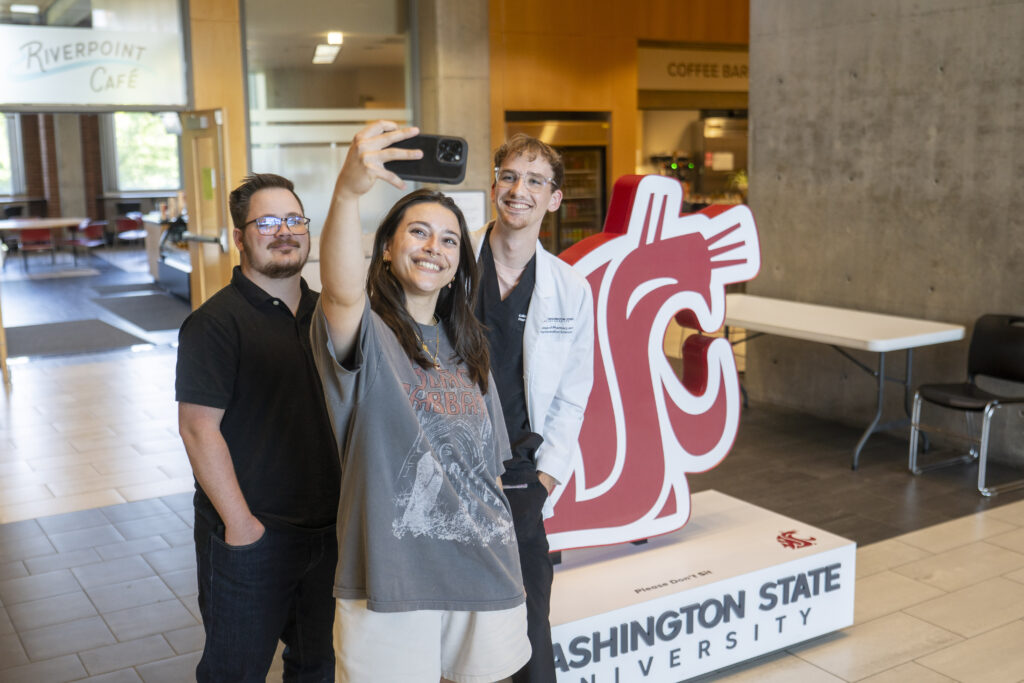 Three people smiling while taking a group selfie in front of a WSU cougar logo installation.