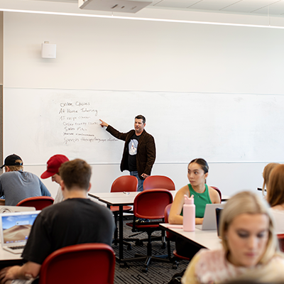 Instructor standing at wall mounted white board with notes written on it, teaching students at their desks