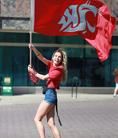 A person wearing a red shirt and blue shorts waves a large red Washington State University flag while holding pom-poms, with a glass-windowed building and another person in the background.