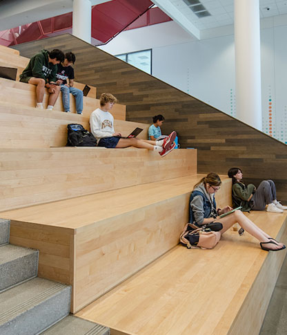 A group of students sitting on wooden bleachers inside a modern building, engaged in activities like reading, using laptops, and talking with one another.