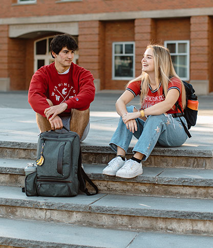 Two people sitting on outdoor steps in front of a brick building. One wears a red 