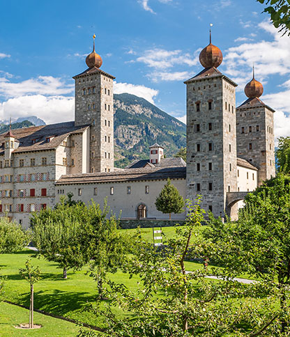 A historic castle with three onion-shaped domes set against a mountain backdrop and blue sky, surrounded by a lush garden with trees and shrubs.