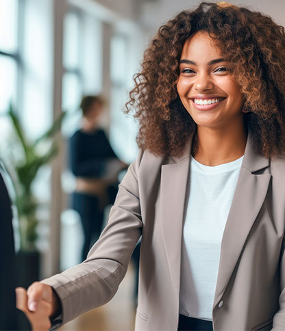 A person with curly hair wearing a light-colored blazer and white shirt extends a hand for a handshake in a professional indoor setting, with another person and plants visible in the background.