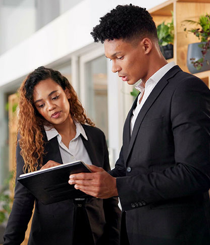 Two individuals in business attire standing indoors, with one person holding a tablet and showing something to the other. Shelves with plants are visible in the background.