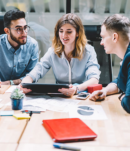 Four people sitting around a table engaged in discussion, with papers showing charts and graphs, a red notebook, sticky notes, a small potted plant, and electronic devices like smartphones and tablets.