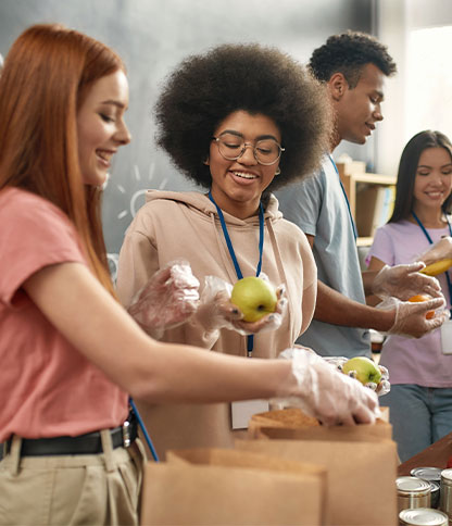 A group of people wearing gloves and name tags pack apples and oranges into brown paper bags in a community service setting, with shelves of supplies in the background.