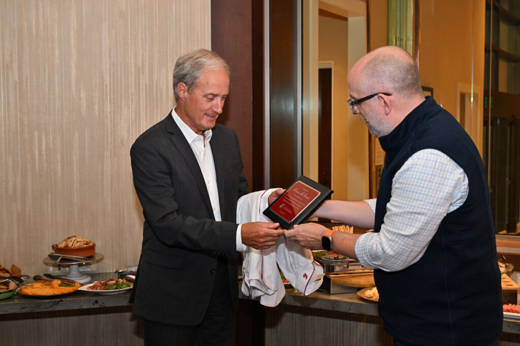 Oliver Meinzer presents an award plaque to Brian McGinnis in recognition of his dedicated service and lasting impact on the School of Hospitality Business Management Advisory Board, with a buffet table in the background.