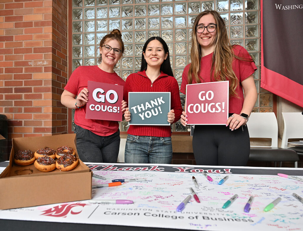 Three students wearing crimson and red hold signs reading “GO COUGS!” and “THANK YOU!” behind a table with a thank-you banner, colorful pens, and a box of chocolate-covered donuts during WSU Giving Day.