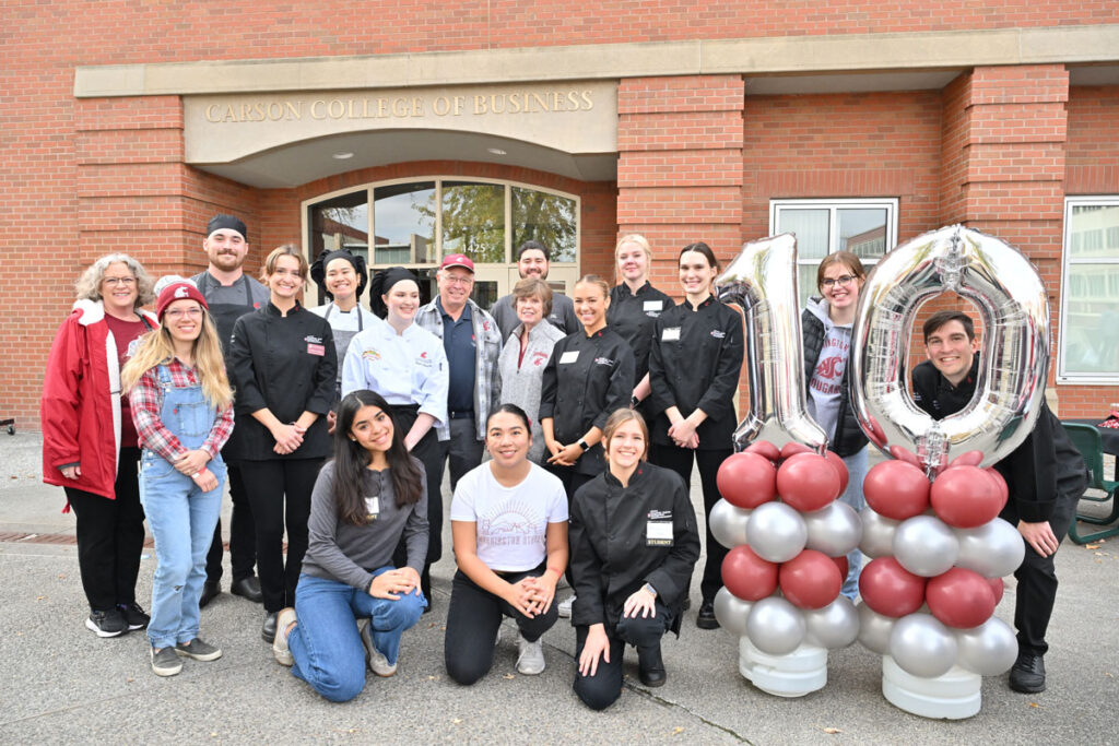 A group poses in front of the Carson College of Business building during WSU Homecoming 2024, standing beside silver balloons forming the number 10 to celebrate the 10th anniversary of the college’s naming.