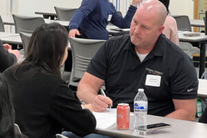 Coach Chuck Evers sits at a table during a coaching session, writing on a sheet of paper with a water bottle and beverage can in front of him.