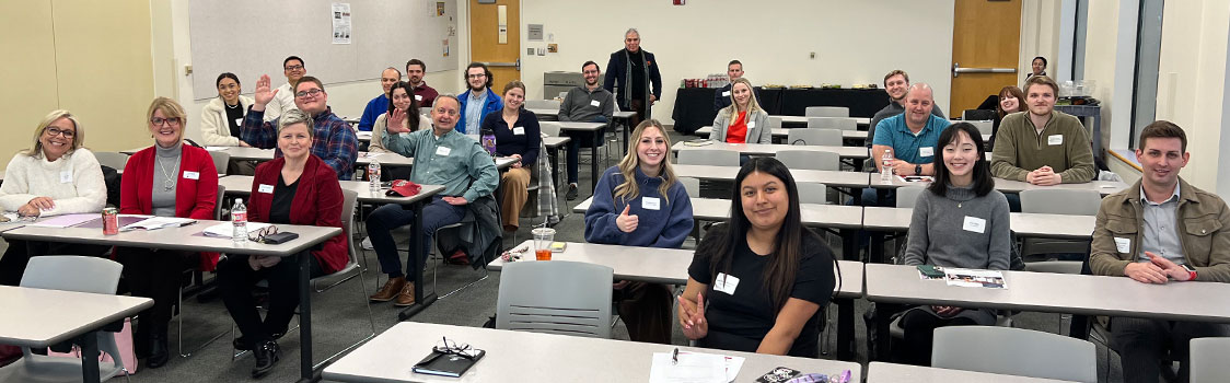 Group of participants seated at classroom-style tables during a Carson Coaching Program session. Name tags, notebooks, and drinks are visible on the tables, with a presenter standing at the back of the room.