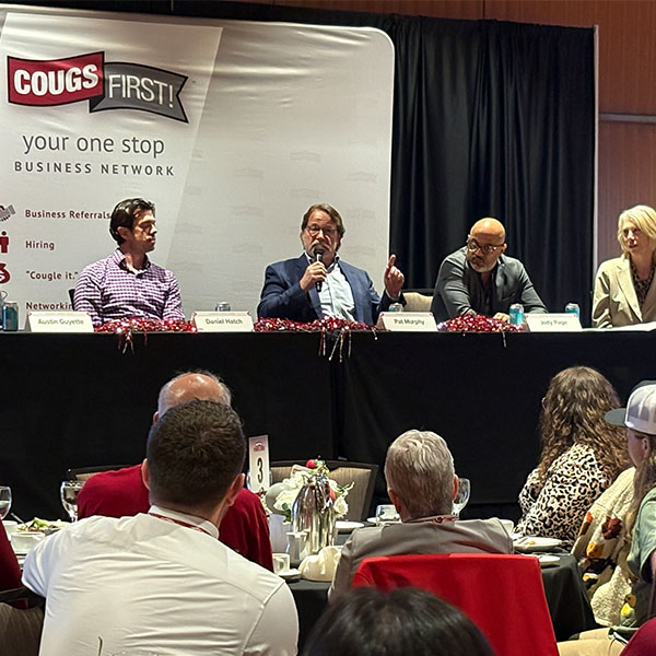 Panel discussion at a CougsFirst! event with five seated speakers and a banner displaying the CougsFirst! logo.