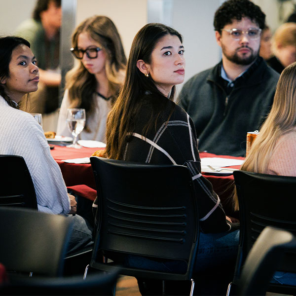 People seated at round tables during an indoor event, with glasses and table settings visible.