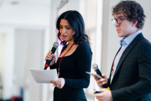 Allison Reyes-Davies and Caden Richard stand side by side, holding microphones and notes while emceeing the Women in Sales event.
