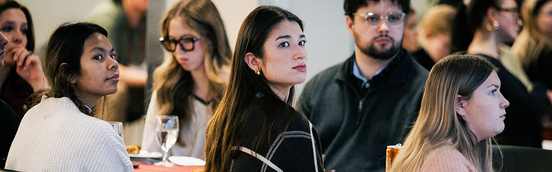 People seated at round tables during an indoor event, with glasses and table settings visible.