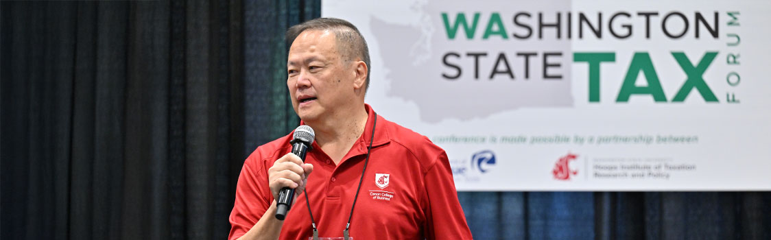 Distinguished Professor Bernard Wong-On-Wing holding a microphone in front of a Washington State Tax Forum banner.