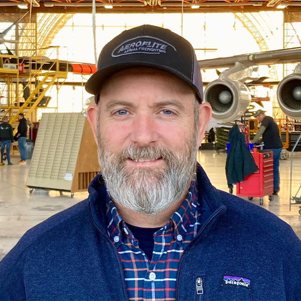 Kelly Anthony standing in an aircraft hangar with two planes and maintenance equipment in the background.