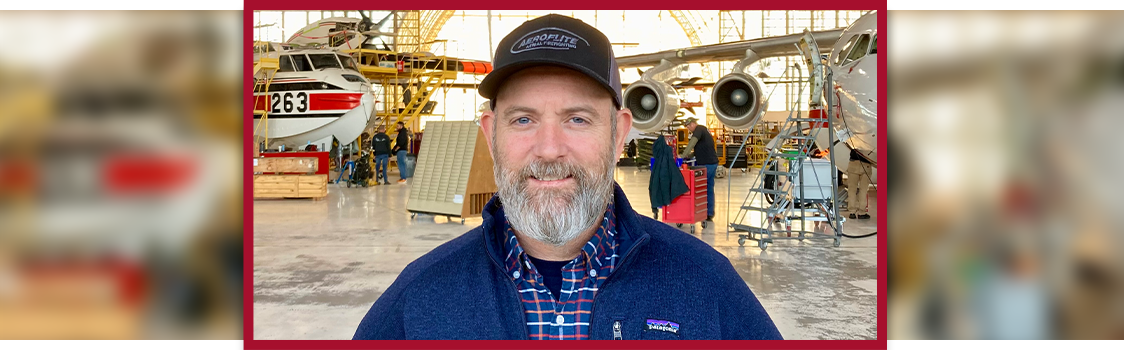 Kelly Anthony standing in an aircraft hangar with two planes and maintenance equipment in the background.