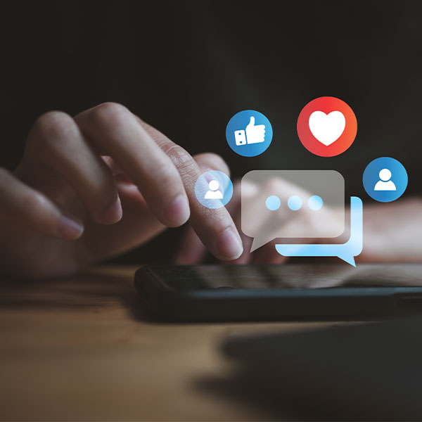 Close-up of a hand interacting with a smartphone, with digital icons for social media engagement—like, heart, chat, and user—floating above the screen.
